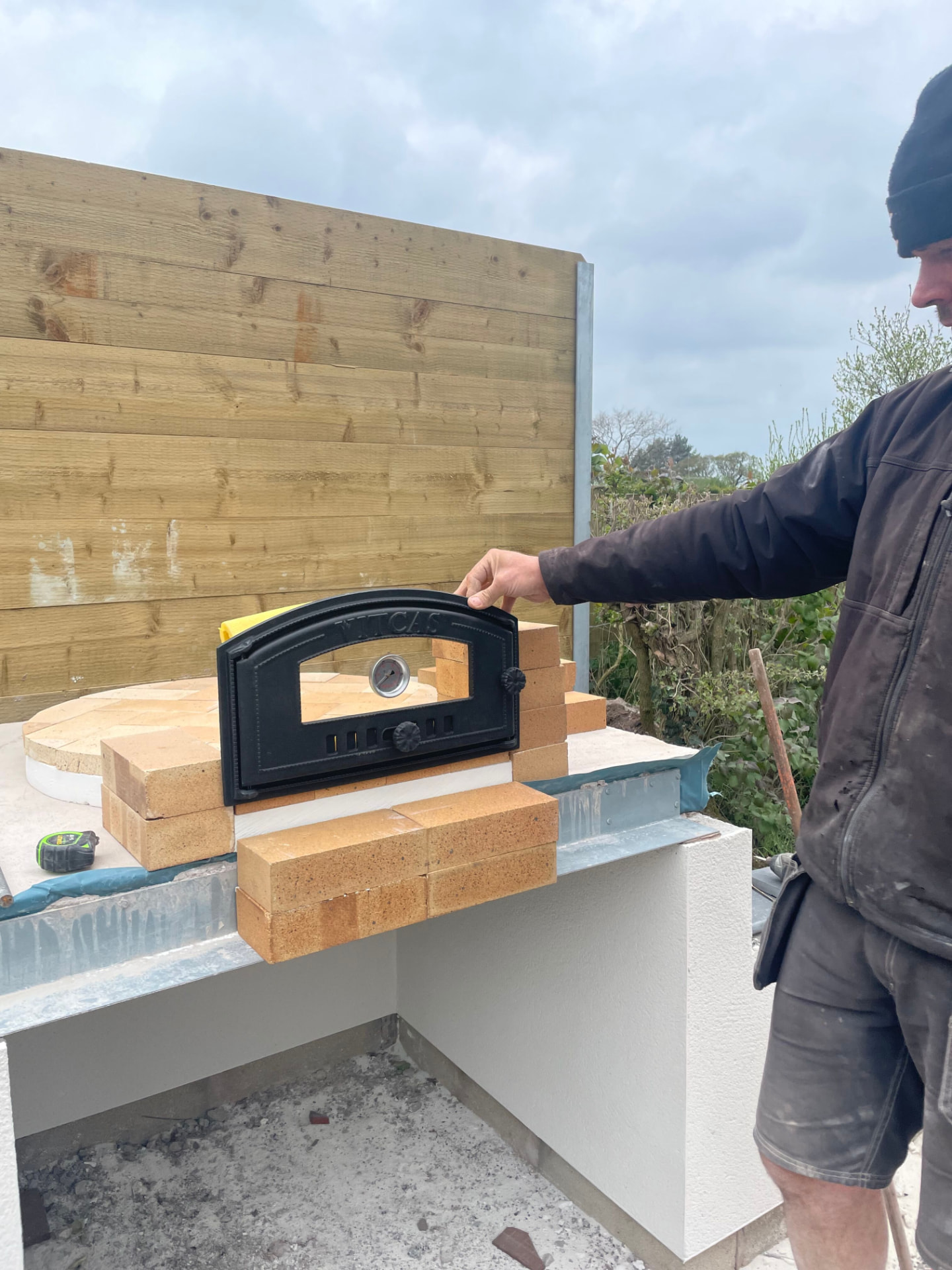 Cast-iron oven door and firebricks being set in place on the pizza oven base