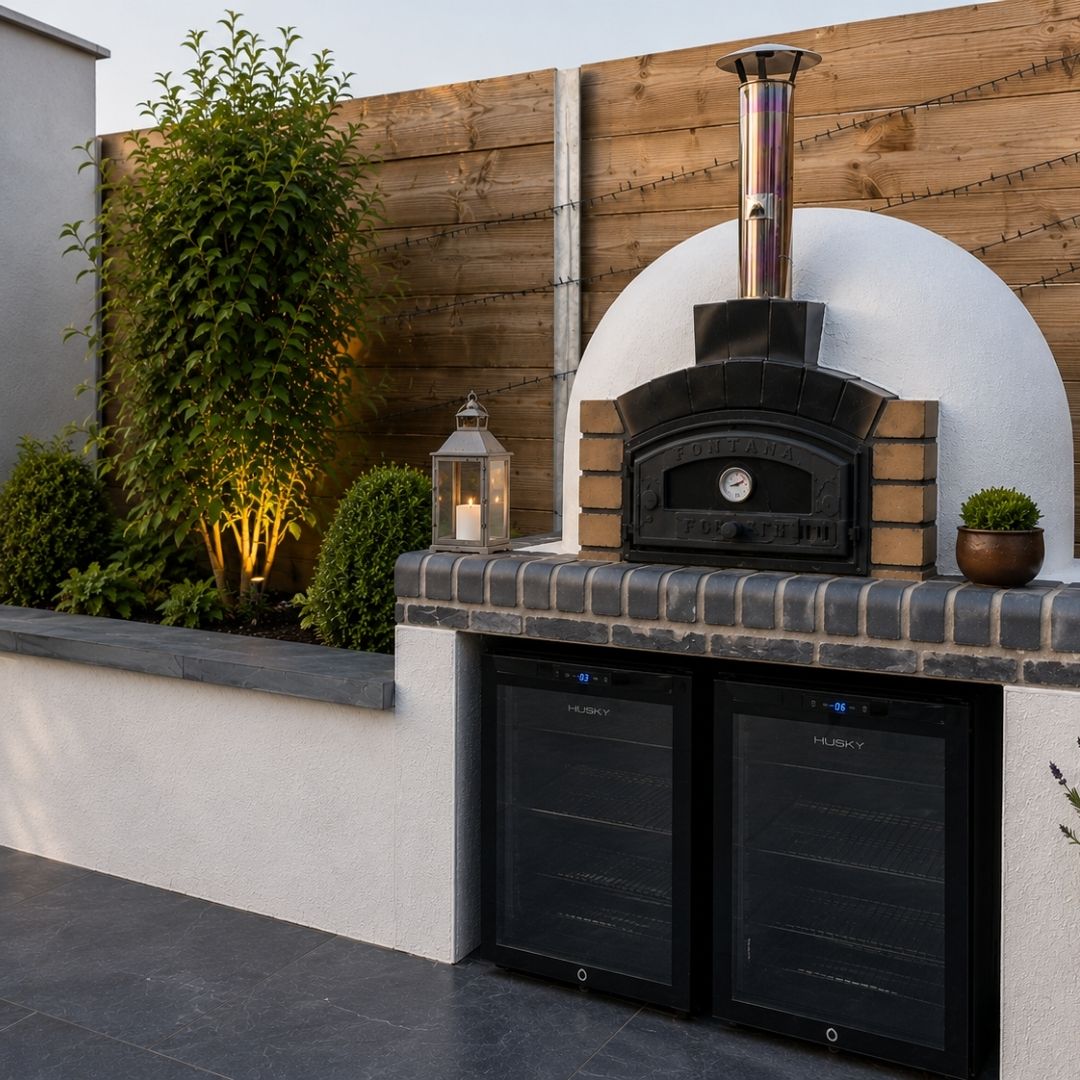 Outdoor kitchen at dusk with lit pizza oven, brick coping, built-in fridges and planting